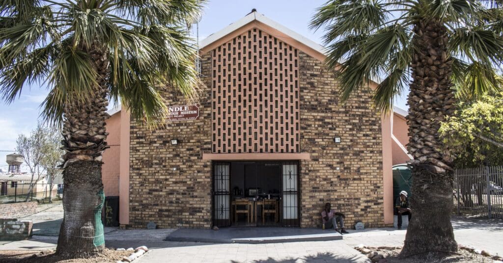 Community museum with brick facade, triangular roof, palm trees, seated individuals near entrance.