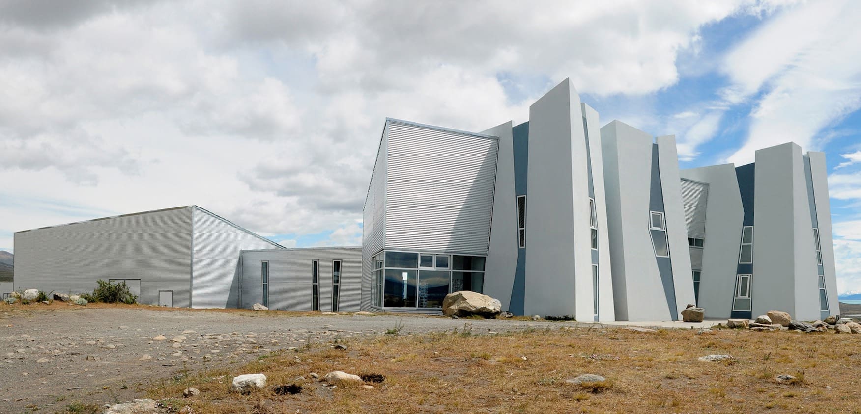 Contemporary angular building with gray metal paneling, tall windows, and scattered rocks under cloudy sky.