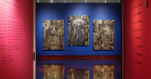 Contemporary art exhibition with three abstract artworks on a blue wall, flanked by text on red walls.