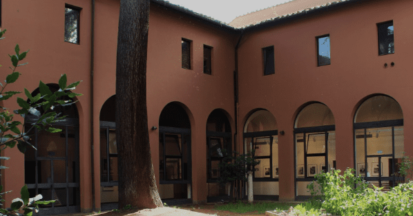 Courtyard with arched doorways, red-brown walls, tall tree trunk, and landscaped greenery.