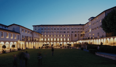 Expansive courtyard surrounded by a lit multi-story building with arched colonnades at dusk.