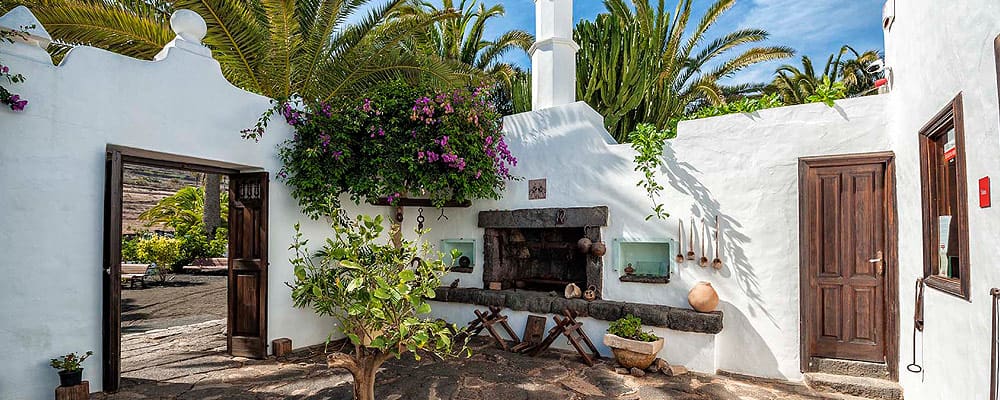 Courtyard with white walls, wooden doors, palm trees, flowering plants, and stone fireplace with pottery.