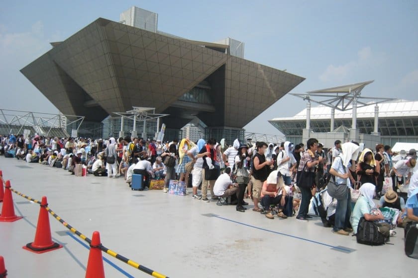 Crowd of people lined up outside a modern geometric building, some providing their own shade.