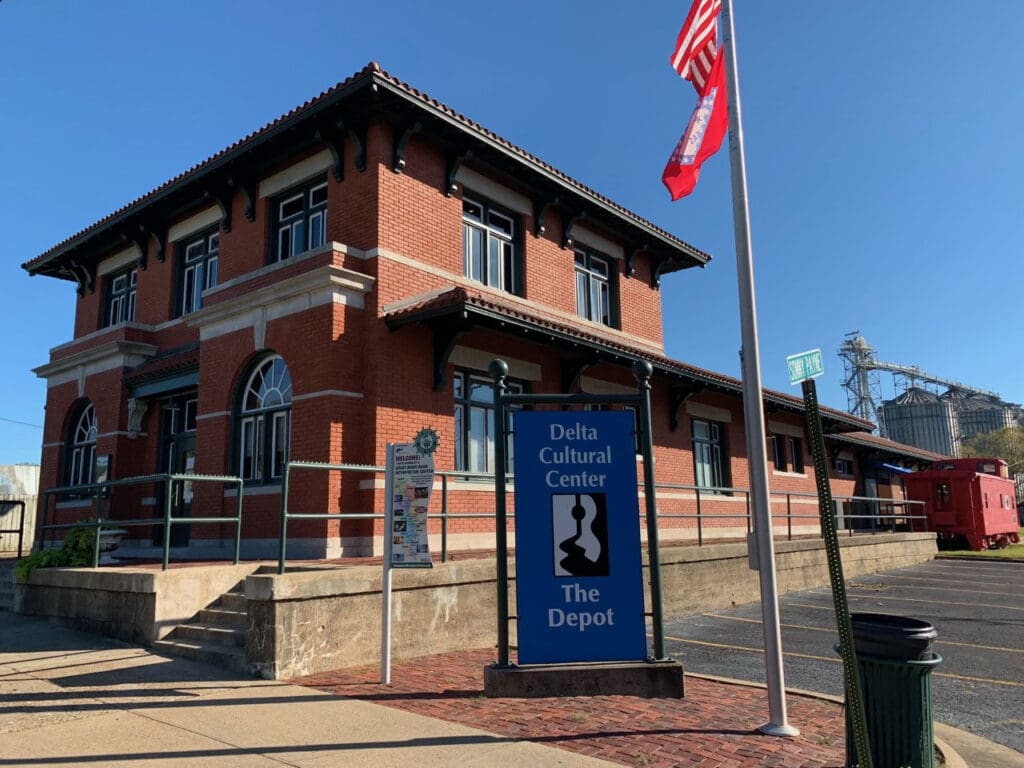 Two-story brick Delta Cultural Center - The Depot with arched windows, flags, and red train car nearby.
