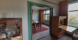 Dining room with central table, chairs, red carpet, large windows, glass cabinet, and wooden desk.