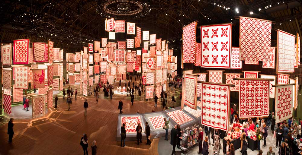 Exhibition hall with red-and-white quilts hanging, visitors observing intricate patterns.