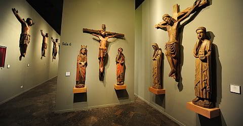 Exhibition hall with religious wooden sculptures on green walls, featuring crucifixion scenes. "Sala 6" sign visible.