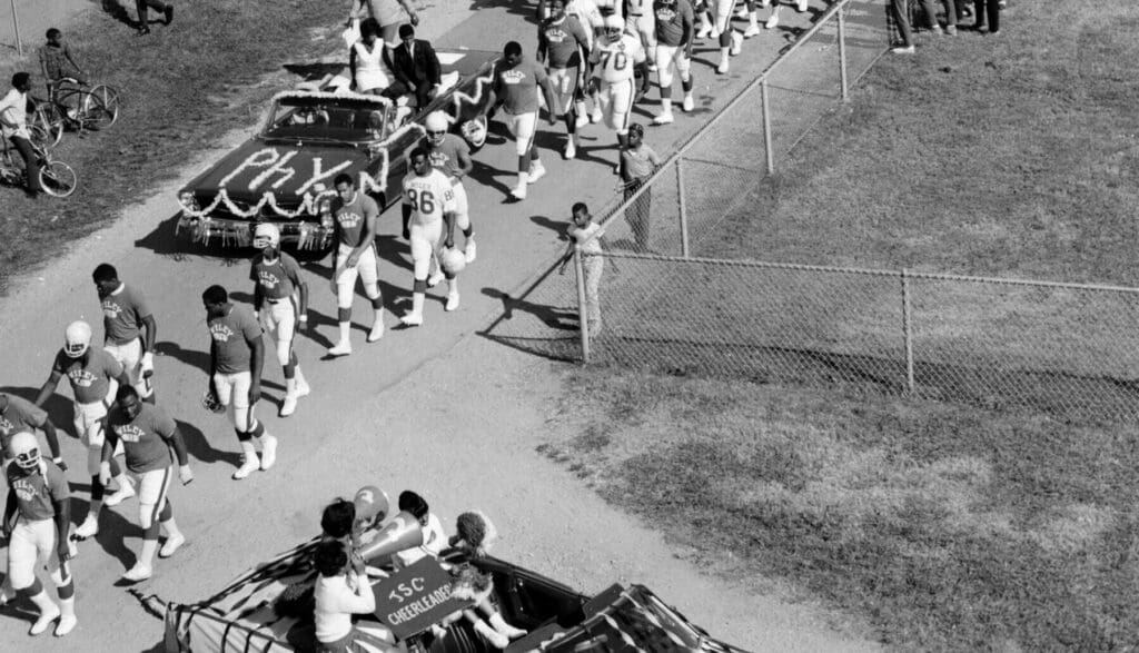 Football players and cheerleaders in a parade with decorated cars and spectators on a dirt path.