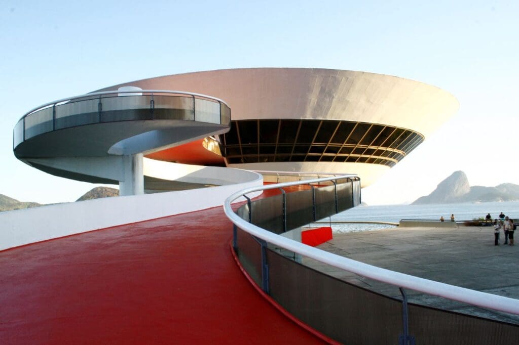 Futuristic building with disc-shaped top, red walkway, ocean views, mountains, and people near water.