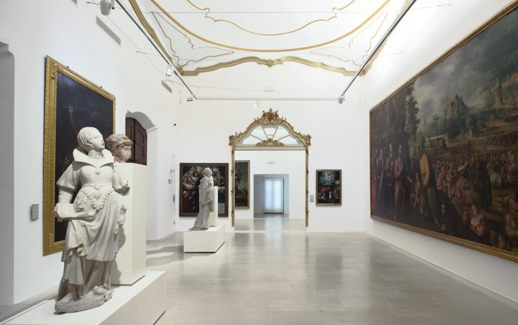 Gallery room with classical sculptures, large framed paintings, ornate ceiling, and polished stone floor.