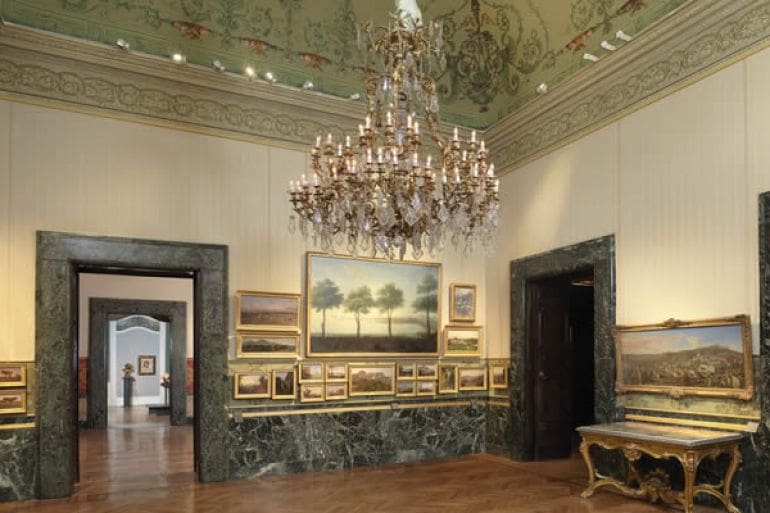 A gallery room with a crystal chandelier, framed paintings, ornate ceiling, and marble doorways