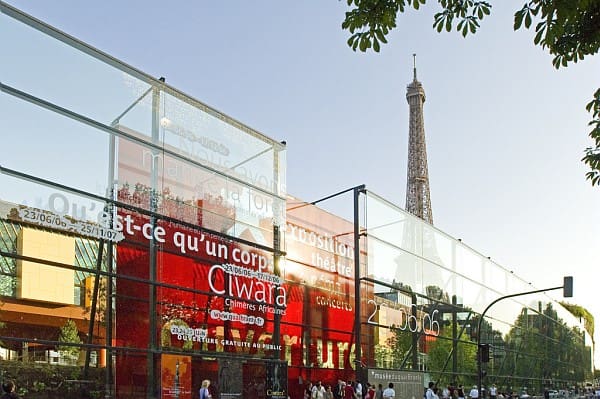 Glass-walled building with event signage, people nearby, and Eiffel Tower in the distant background.