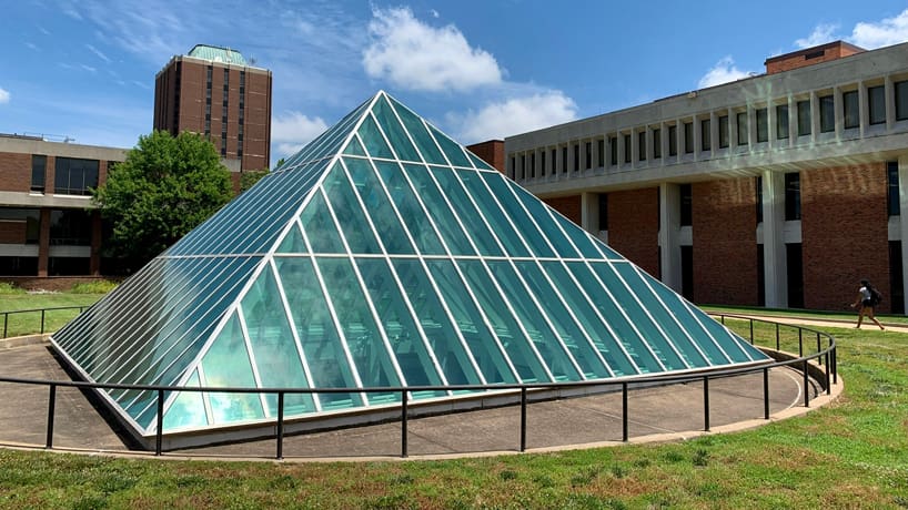 Glass pyramid with metal railing in grassy area, surrounded by brick buildings with rectangular windows