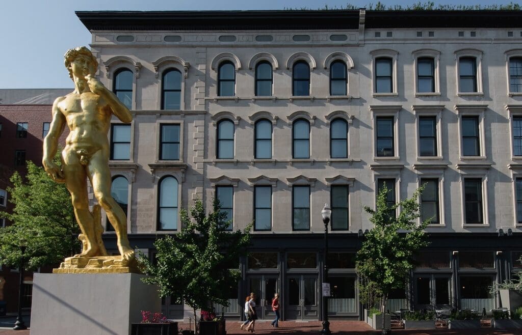 Golden nude male statue in front of multi-story building with arched windows, trees, streetlamp, pedestrians.
