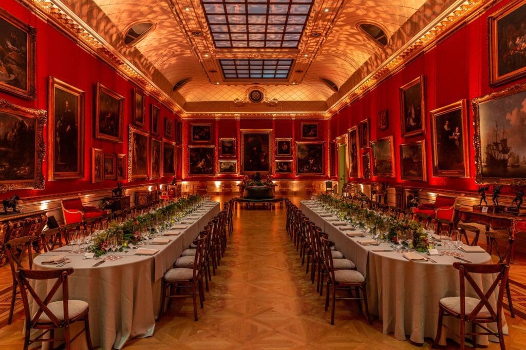 Grand dining hall with high ceilings, ornate lighting, red walls, framed paintings, and banquet tables.