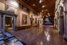 A spacious grand hall with chandeliers, wood ceilings, polished stone flooring, and classical paintings.
