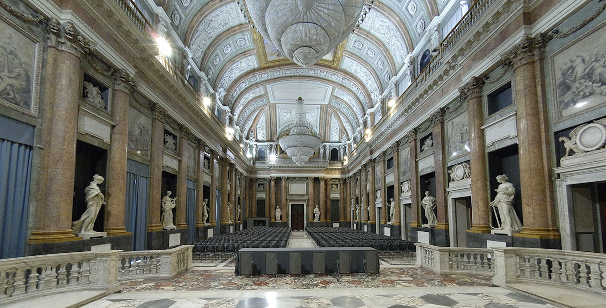 Formal grand hall with ornate architecture, marble columns, statues, chandeliers, and rows of seating