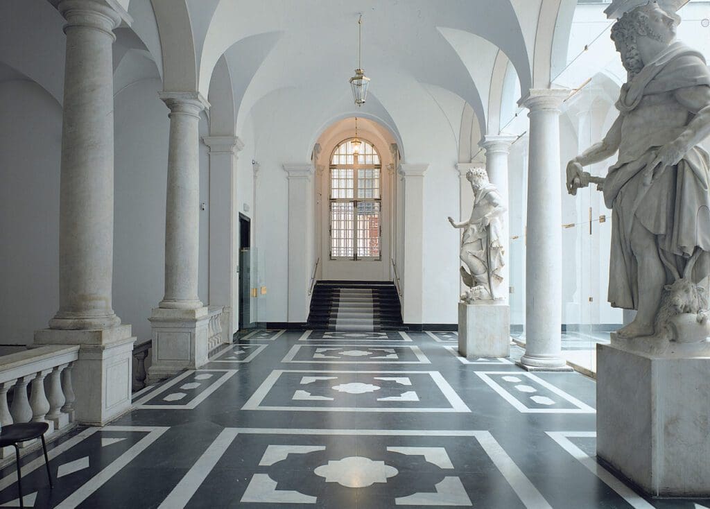Grand interior hallway with high arched ceilings, marble columns, statues, and intricate flooring.