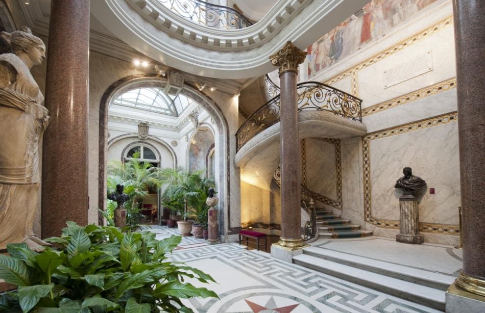 Grand interior with marble columns, decorative staircase, skylight, statues, plants, and detailed mosaics.