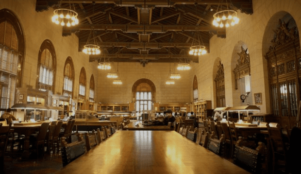 Grand library reading room with high ceilings, chandeliers, arched windows, tables, chairs, and bookshelves.