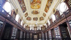 Interior of a grand library with high wooden bookshelves and colorful frescoes on the ornate ceiling.