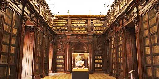 Grand library with wooden shelves, ornate balcony, and central lit display case showcasing an item.
