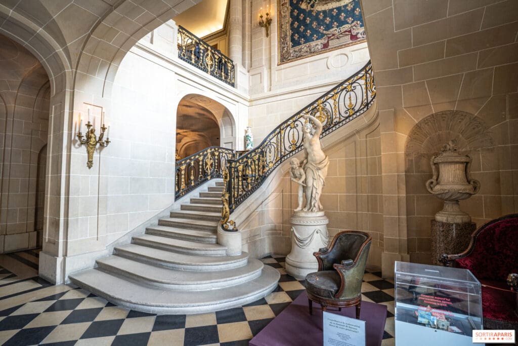 Grand marble staircase with ornate golden railings, statue, tapestries, and a checkered floor in elegant room