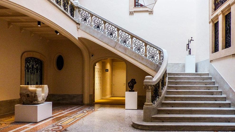 Grand staircase with ornate railings, sculptures on pedestals, arched doorway, and intricate tiled floor