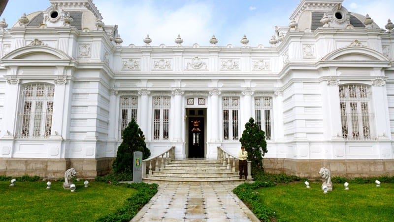 Grand white neoclassical building with a staircase entrance, statues, and trimmed greenery.
