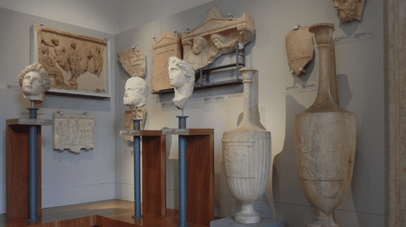 Museum exhibit of classical Greco-Roman sculptures, including marble busts, urns, and relief panels