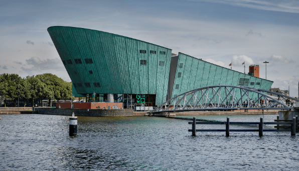 Green-tinted, angular building with pedestrian bridge over water, surrounded by trees and buildings.
