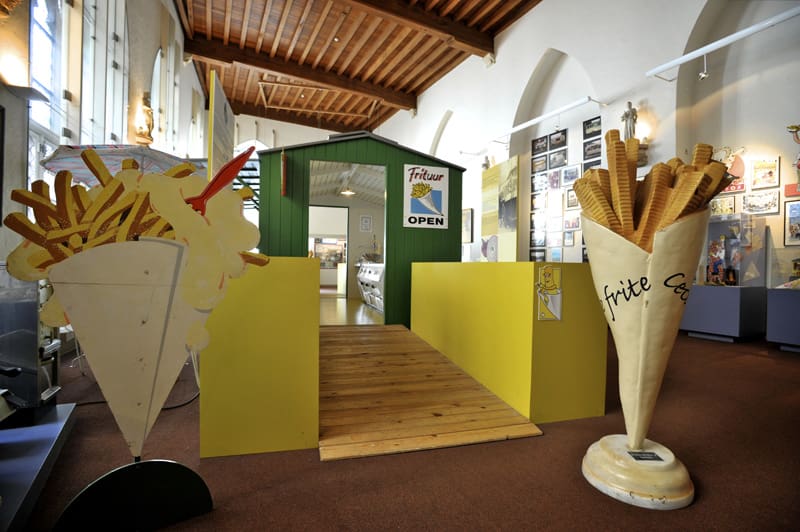 Green kiosk inside a museum labeled "Frituur OPEN," flanked by large cone-shaped fry displays.