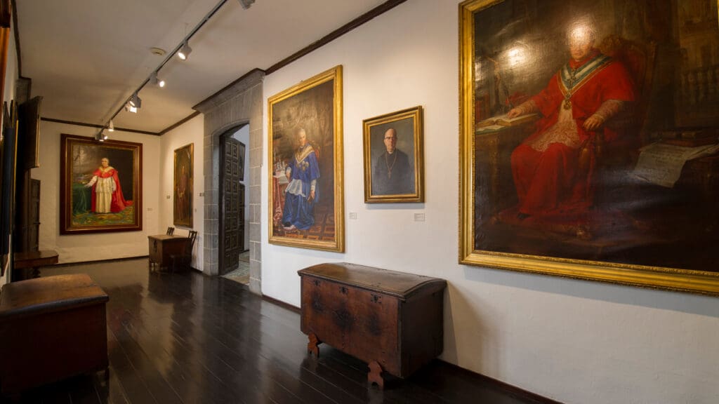 Hallway gallery with large framed portraits of figures in robes, dark wood floors, and wooden chests.