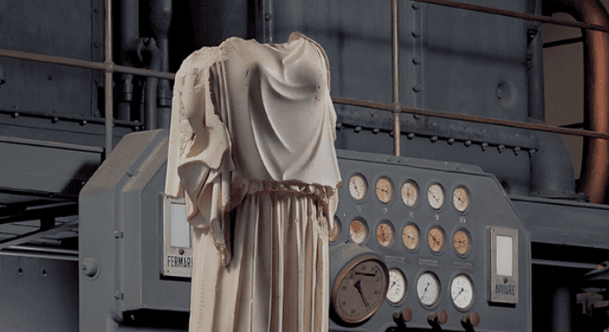 Statue without head or arms in front of industrial control panel with pressure gauges and meters