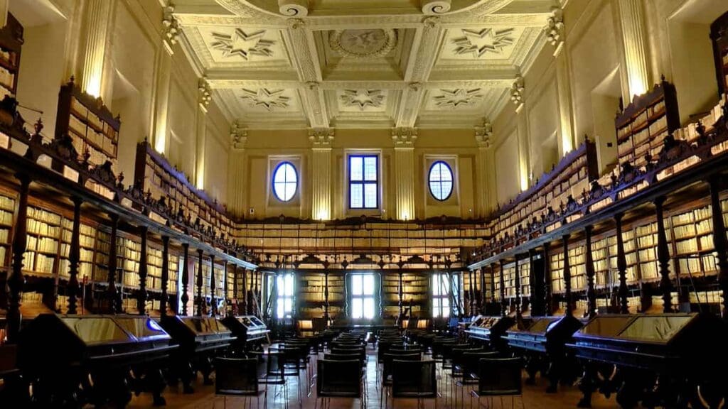 High-ceiling library with decorative moldings, book-filled shelves, desks, chairs, and large windows.