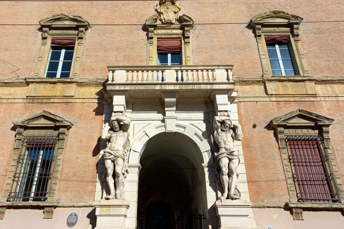 Historic building with ornate arched entrance, scultped male figures, detailed stonework, and balcony