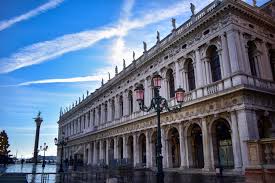 Historic building with arched windows, lamp posts, tall column, under a blue sky with contrails.