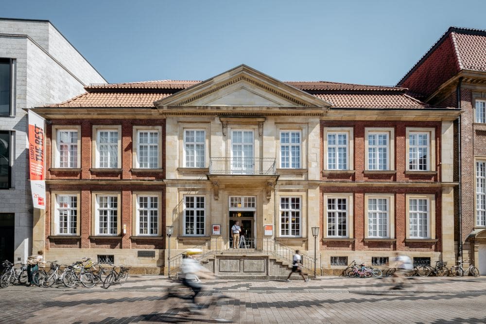 Historic two-story building with large windows, triangular pediment, bicycles, pedestrians, and cyclists.