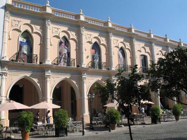 Two-story historic building with arched windows, cafe seating, and umbrellas on a sunny day.