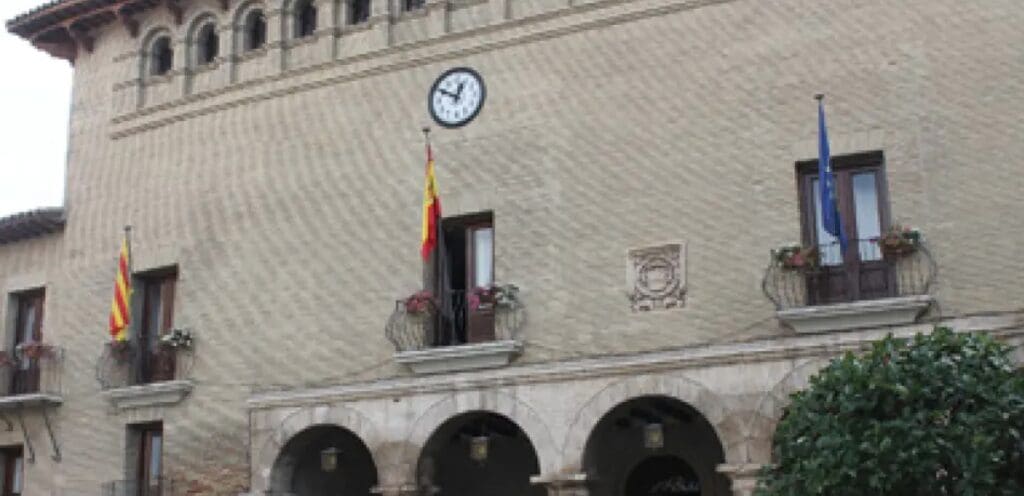 Historic building facade with clock, arched windows, three flags, balconies with flowers, arched doorways.
