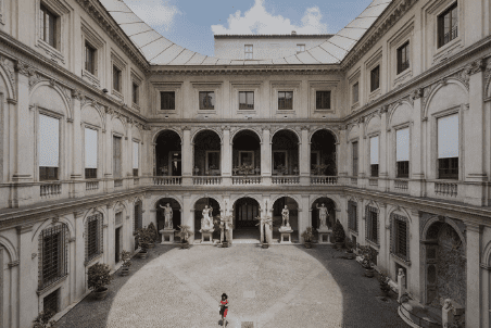 Person in a cobblestone courtyard of a historic multi-story building with statues and arched windows