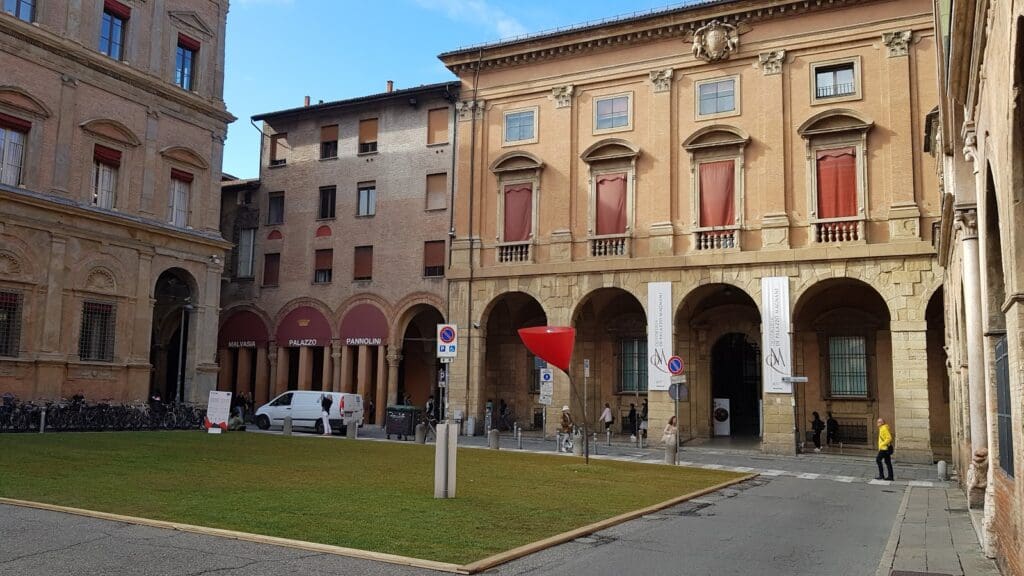 Historic building complex with arched windows, courtyard lawn, signs, people, white van, clear sky.