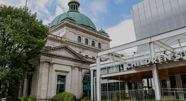 Historic building with green dome, modern structure labeled "Children's," greenery, partly cloudy sky.