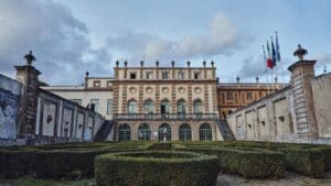 Historic building with ornate façade, arched windows, hedge garden, stone staircases, and flagpoles.