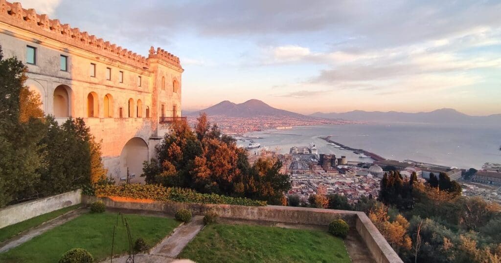 Historic building with arched windows overlooking a city, coastline, bay, and mountains under a partly cloudy sky.
