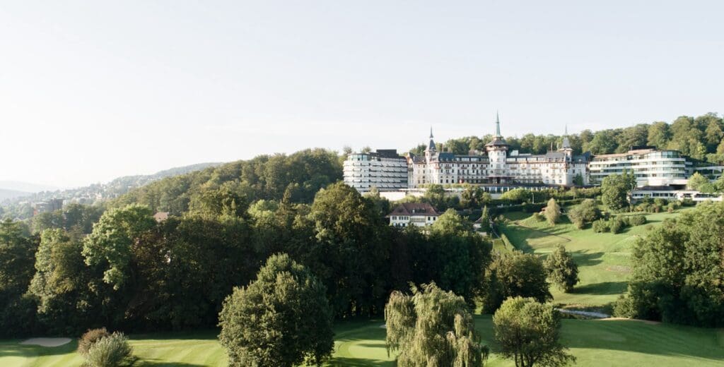 Scenic view of historic building with towers, modern extensions, lush trees, lawns, and hilly background.