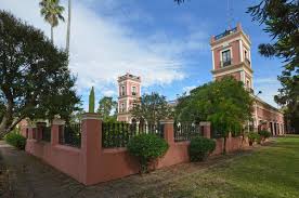 Historic building with two towers, red fence, lush greenery, and clear blue sky in background.