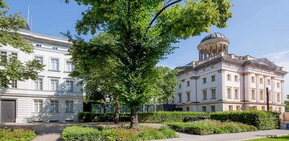 Two historic buildings, a white three-story and an ornate structure with a dome, in a lush setting.