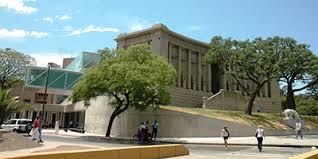 Historic building with classical architecture, trees, modern structures, and pedestrians on pathway.