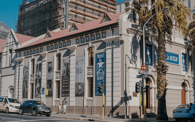 Historic building with red roof housing District Six Museum, posters, vehicles, palm trees, and construction.